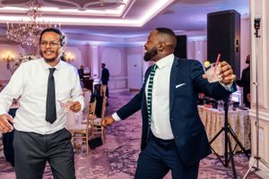 Two wedding guests enthusiastically dancing together in a luxurious ballroom with pink lighting and chandeliers.