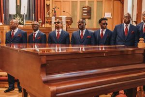 Groomsmen in navy blue suits stand together near a grand piano during a wedding ceremony.
