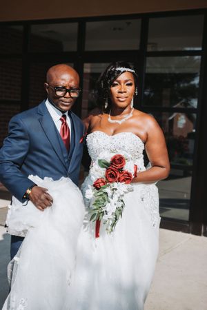 A couple poses in formal wedding attire with red roses outdoors.