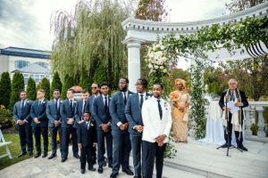 Groomsmen in navy blue suits and one in white stand in formation at a wedding ceremony under a columned arch.