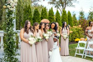 Bridesmaids in matching blush pink dresses pose with the bride holding white bouquets at an outdoor garden venue.