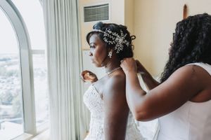 A bride in a strapless wedding dress gets ready near a large arched window with city views.