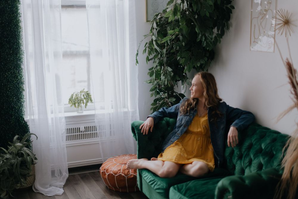 A woman in a yellow dress relaxes on an emerald green velvet couch surrounded by plants.