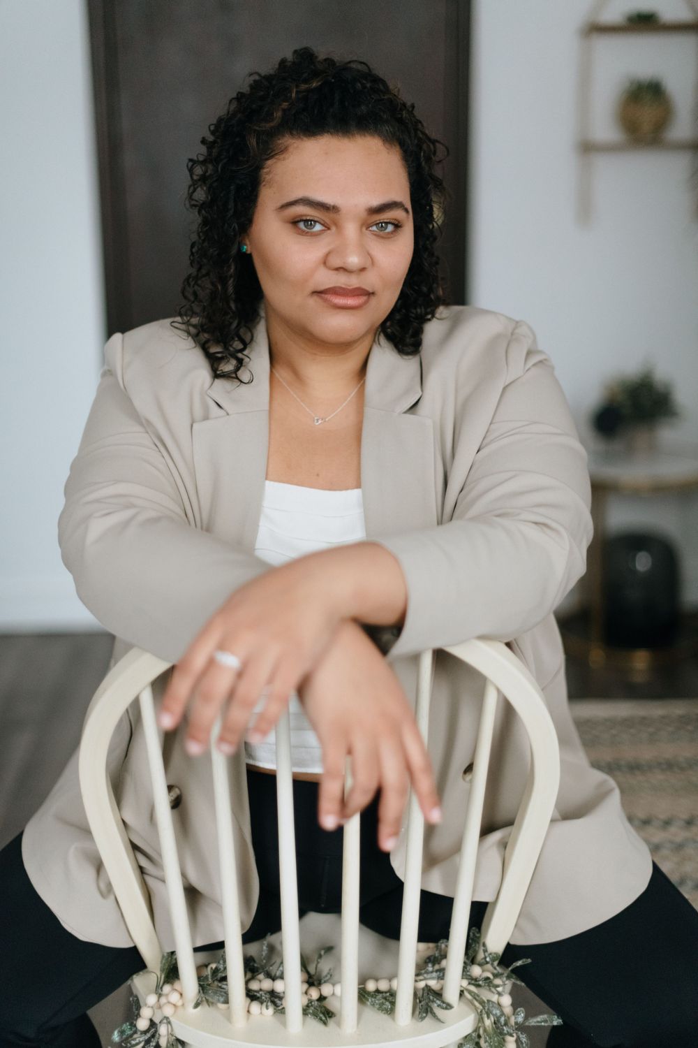 A woman in a beige blazer sits confidently on a white chair in a modern interior.