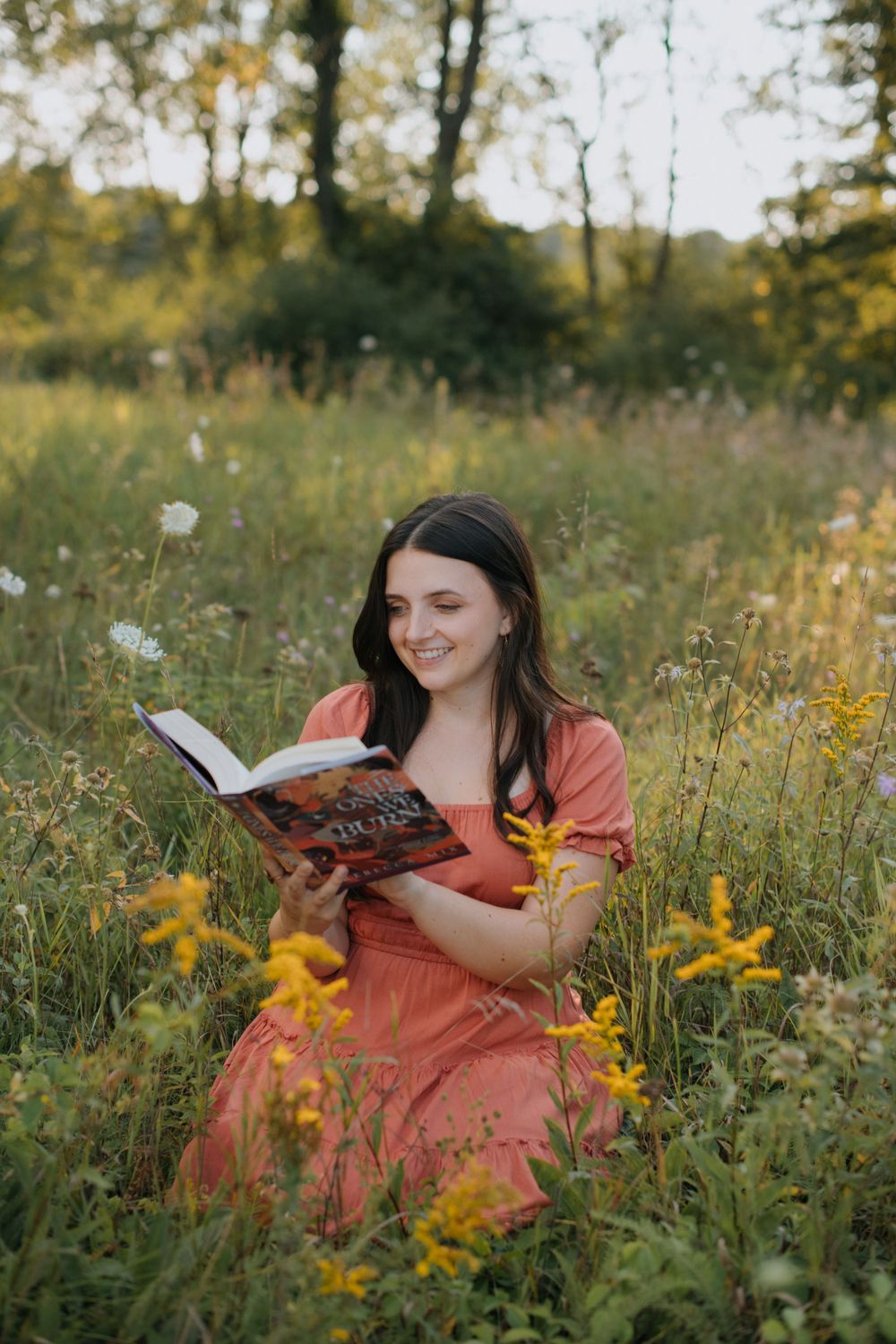 A woman in a coral dress reads a book while sitting in a wildflower meadow at sunset.