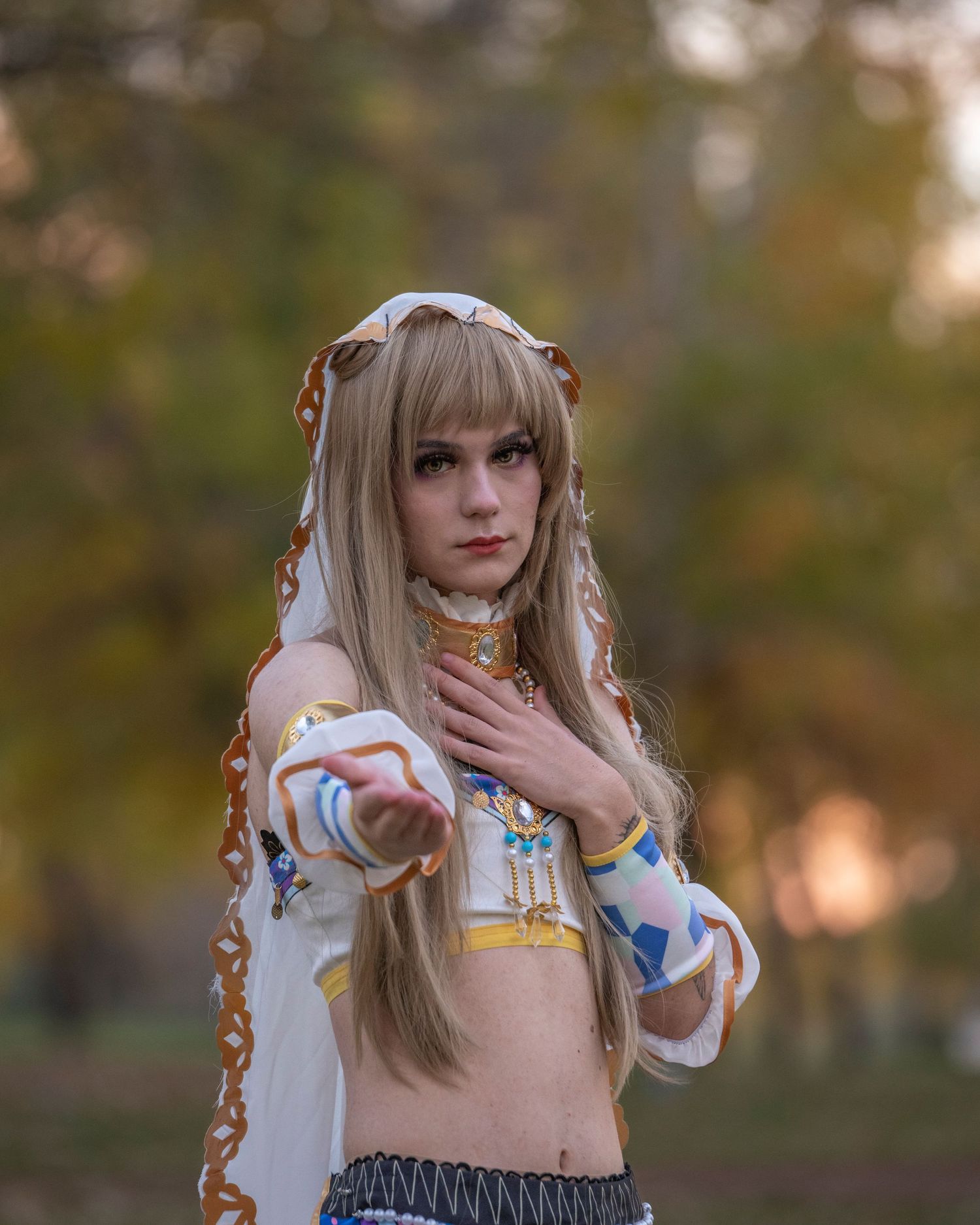 A person in a white costume with colorful details poses outdoors in an autumn setting.