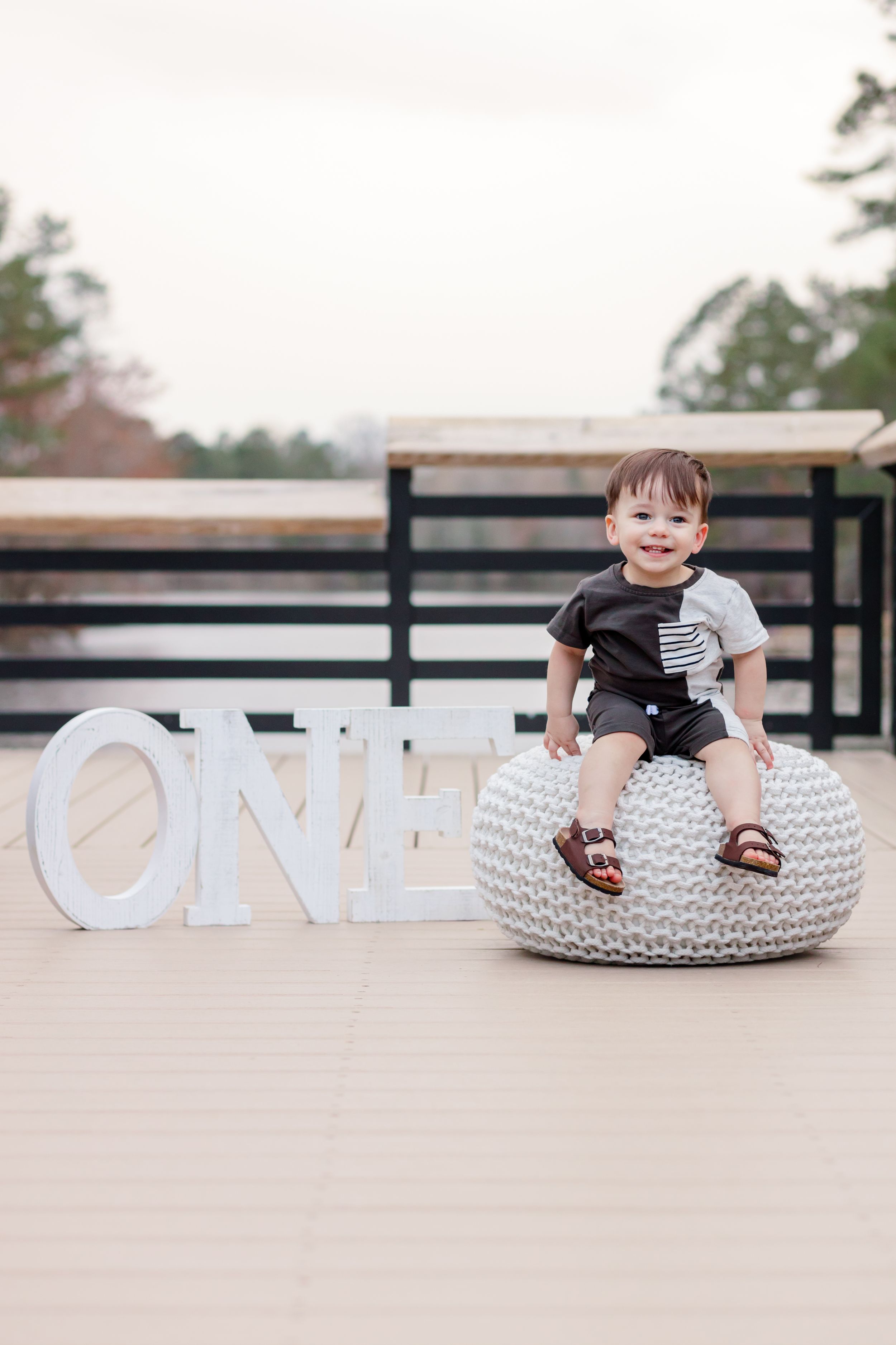 First birthday setup with white knit pouf and 'ONE' letters on outdoor deck.