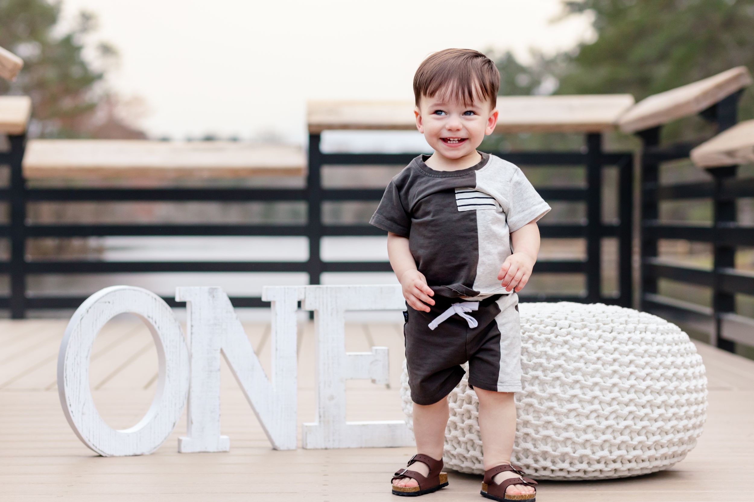 Candid first birthday moment with white knit pouf and 'ONE' letters on wooden deck.