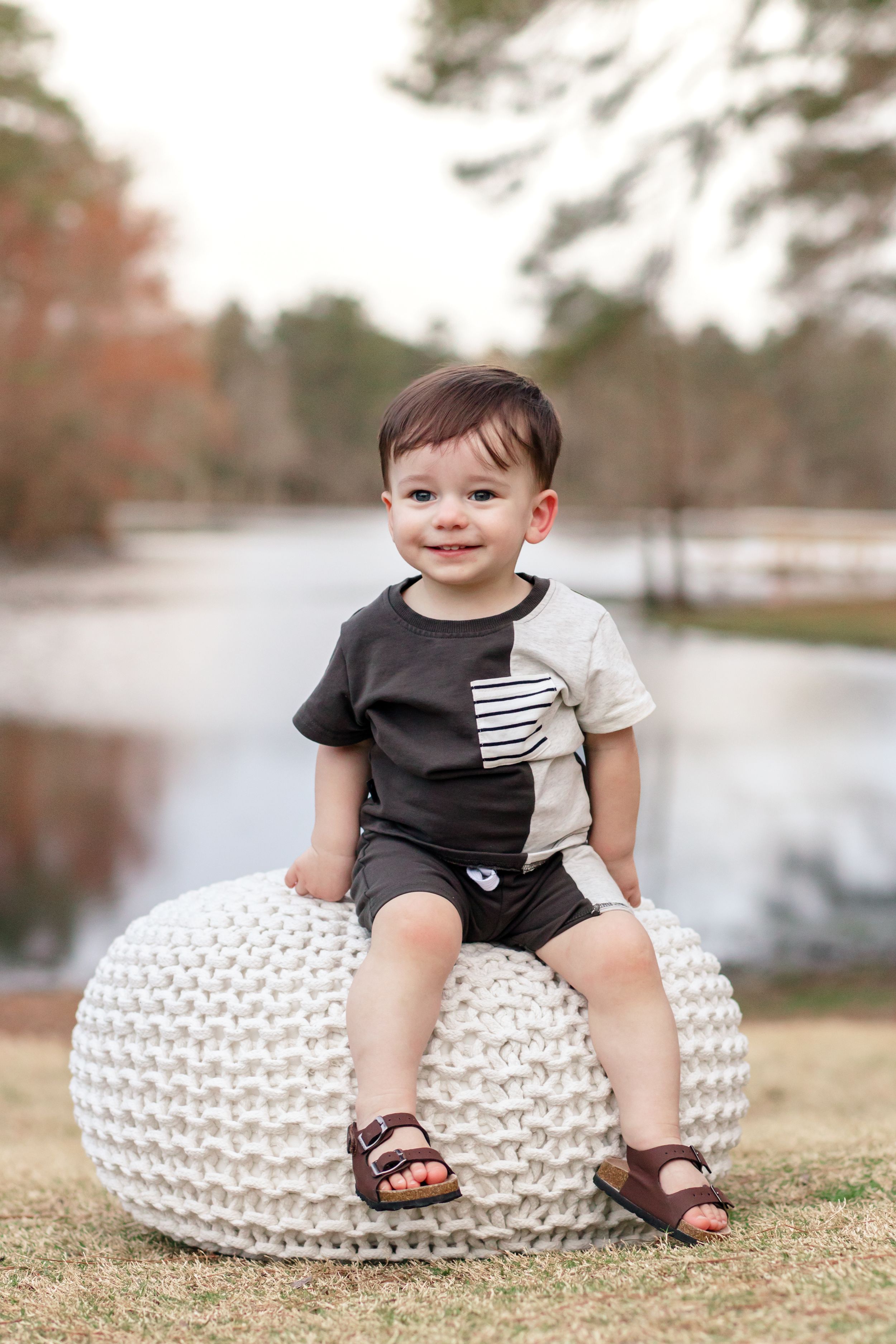 Outdoor first birthday portrait on white knit pouf with lake in background.