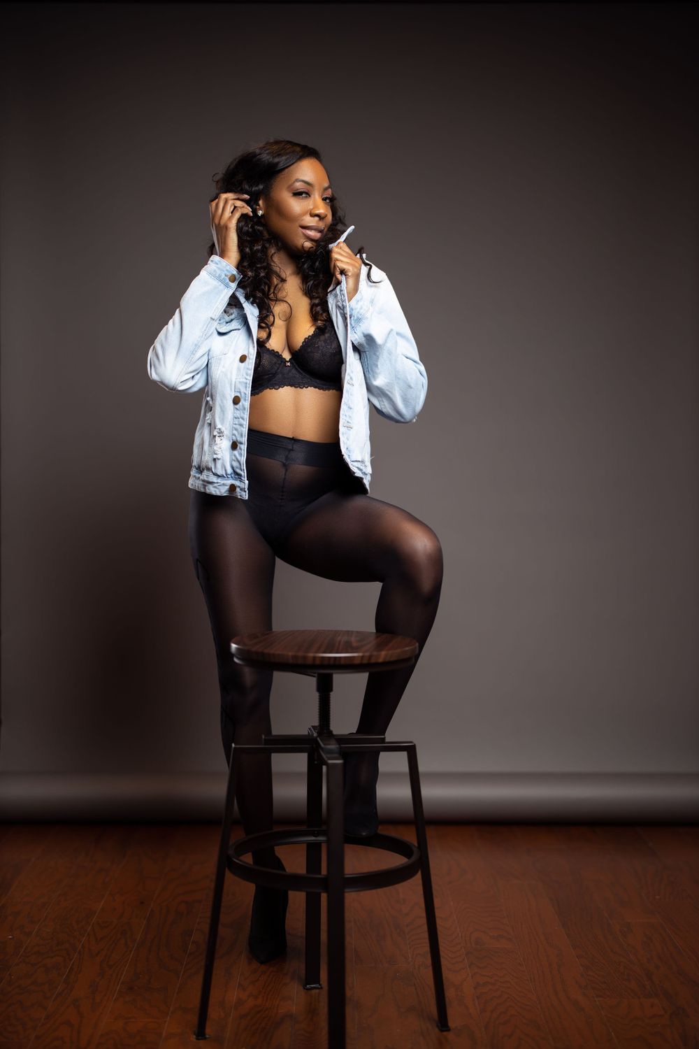 Model in denim jacket poses on barstool wearing black lingerie and sheer tights.