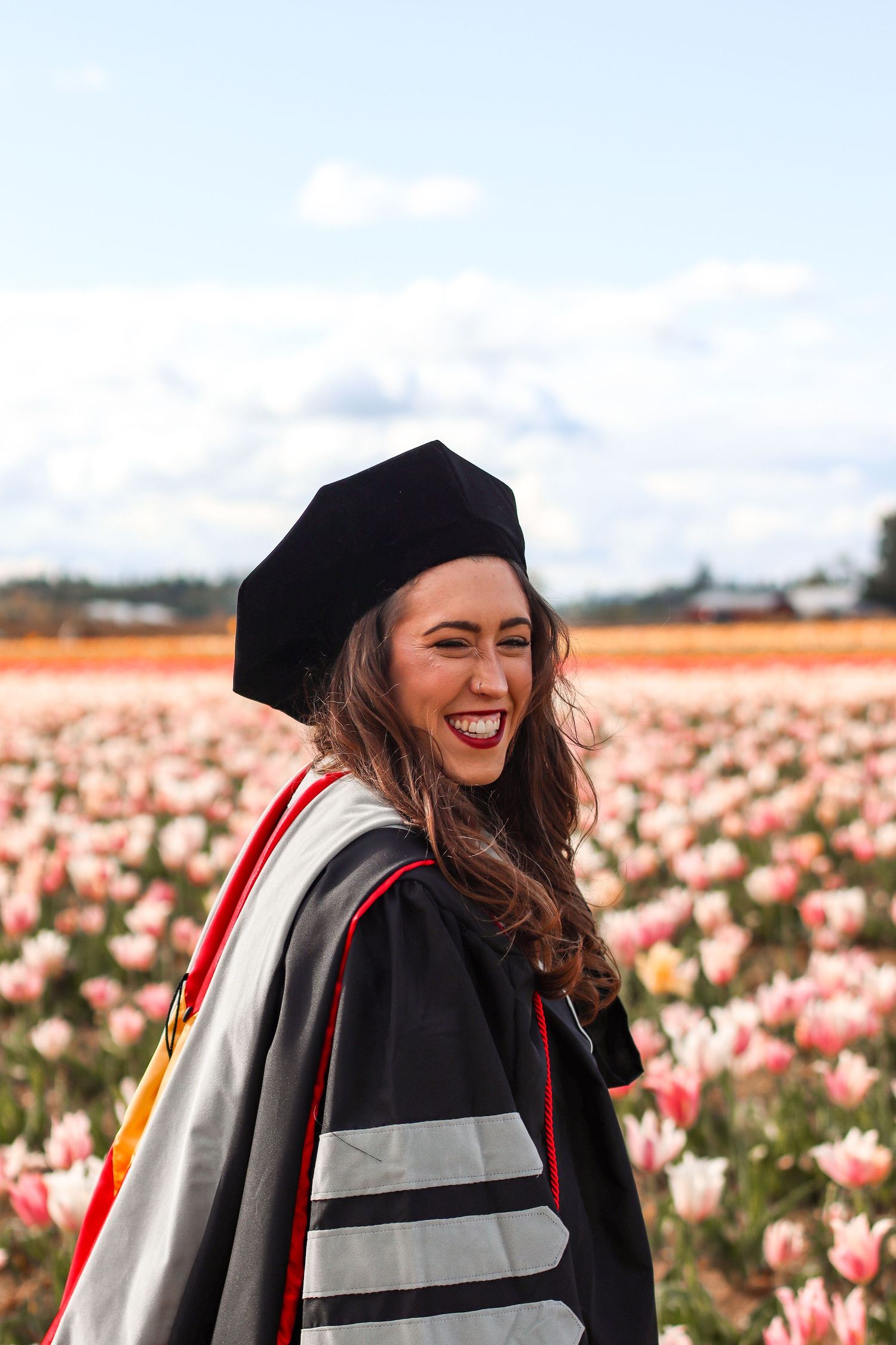Graduate in academic regalia laughs joyfully in front of a vibrant tulip field on sunny graduation day.