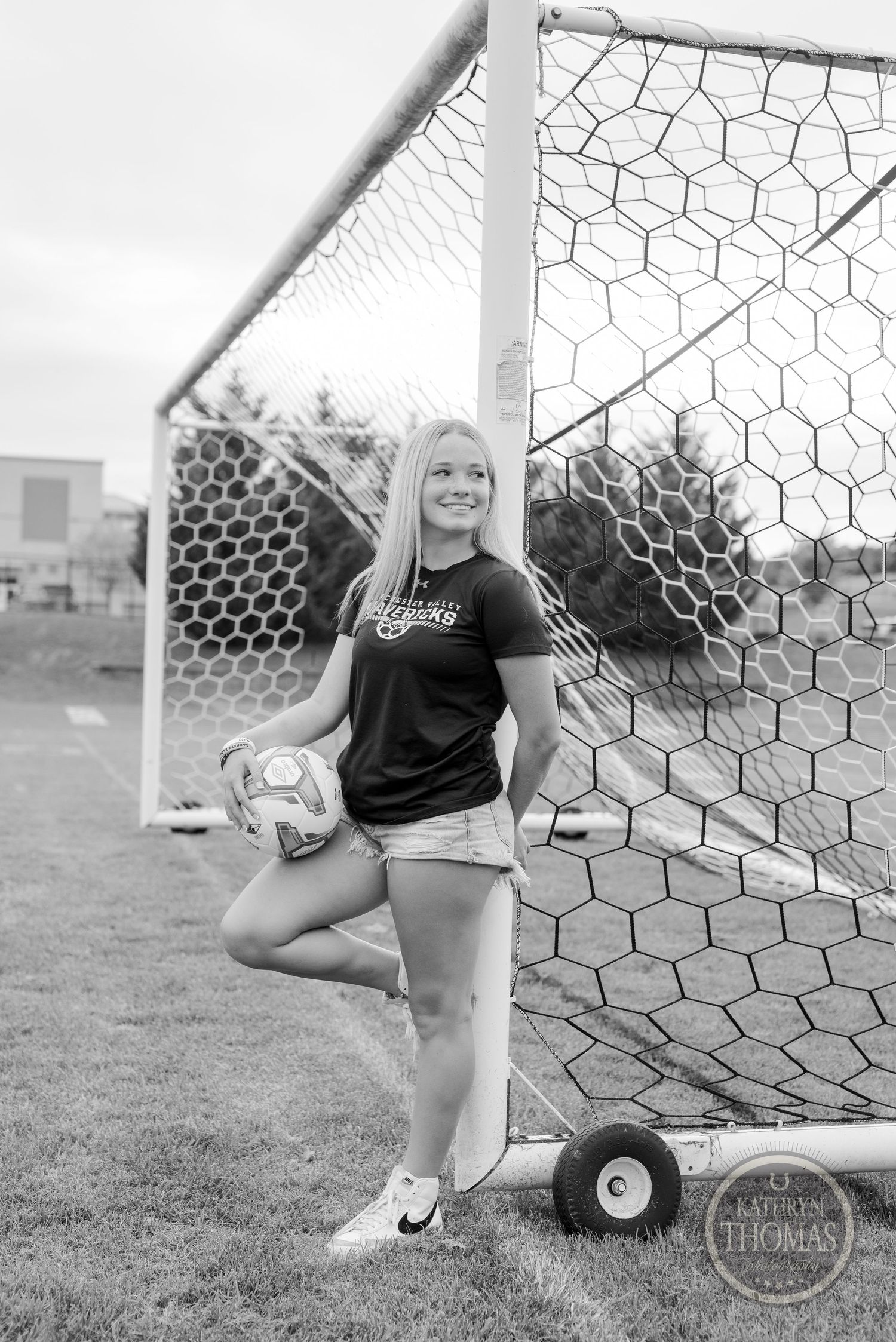 Soccer player in navy uniform posing with ball by goal post in black and white.