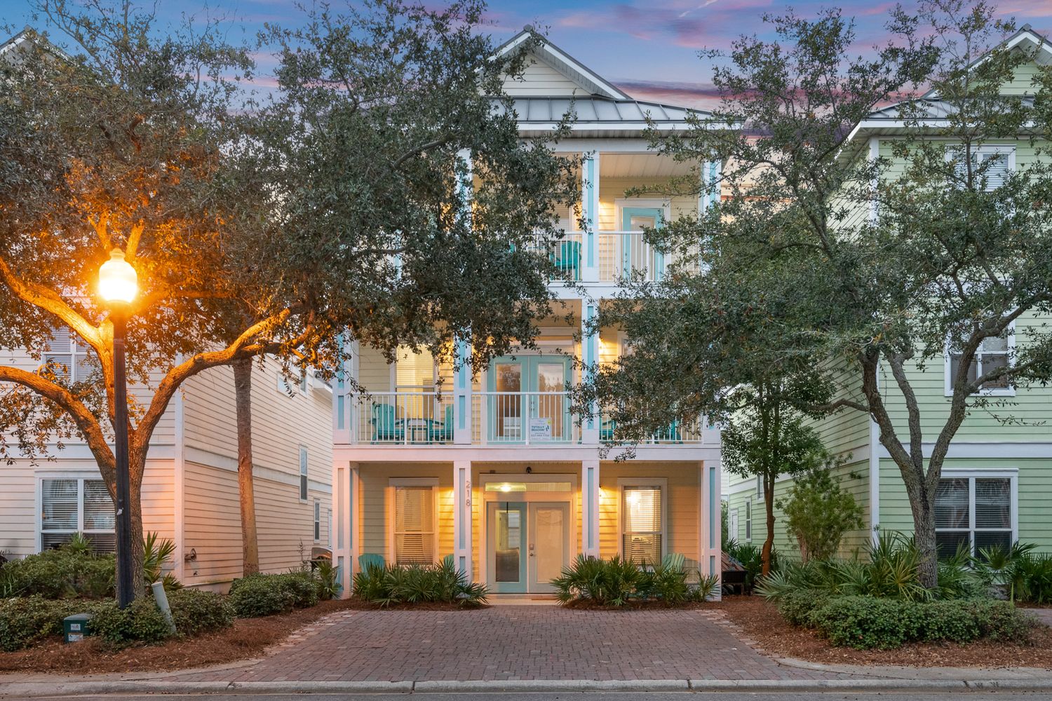 Elegant three-story coastal home with white columns and balconies surrounded by lush landscaping at dusk.