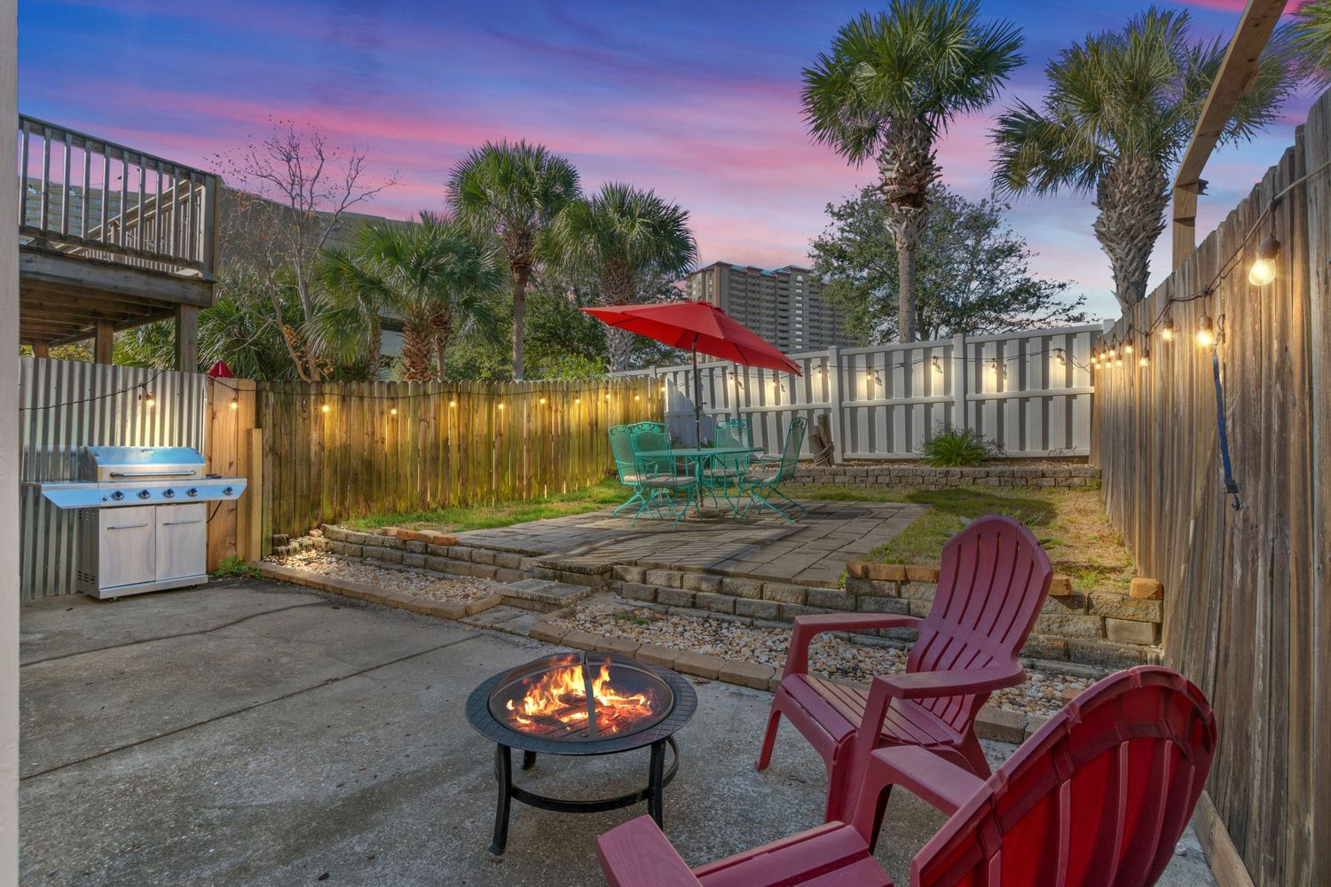 Cozy backyard patio area with fire pit, red Adirondack chairs and string lights at dusk.