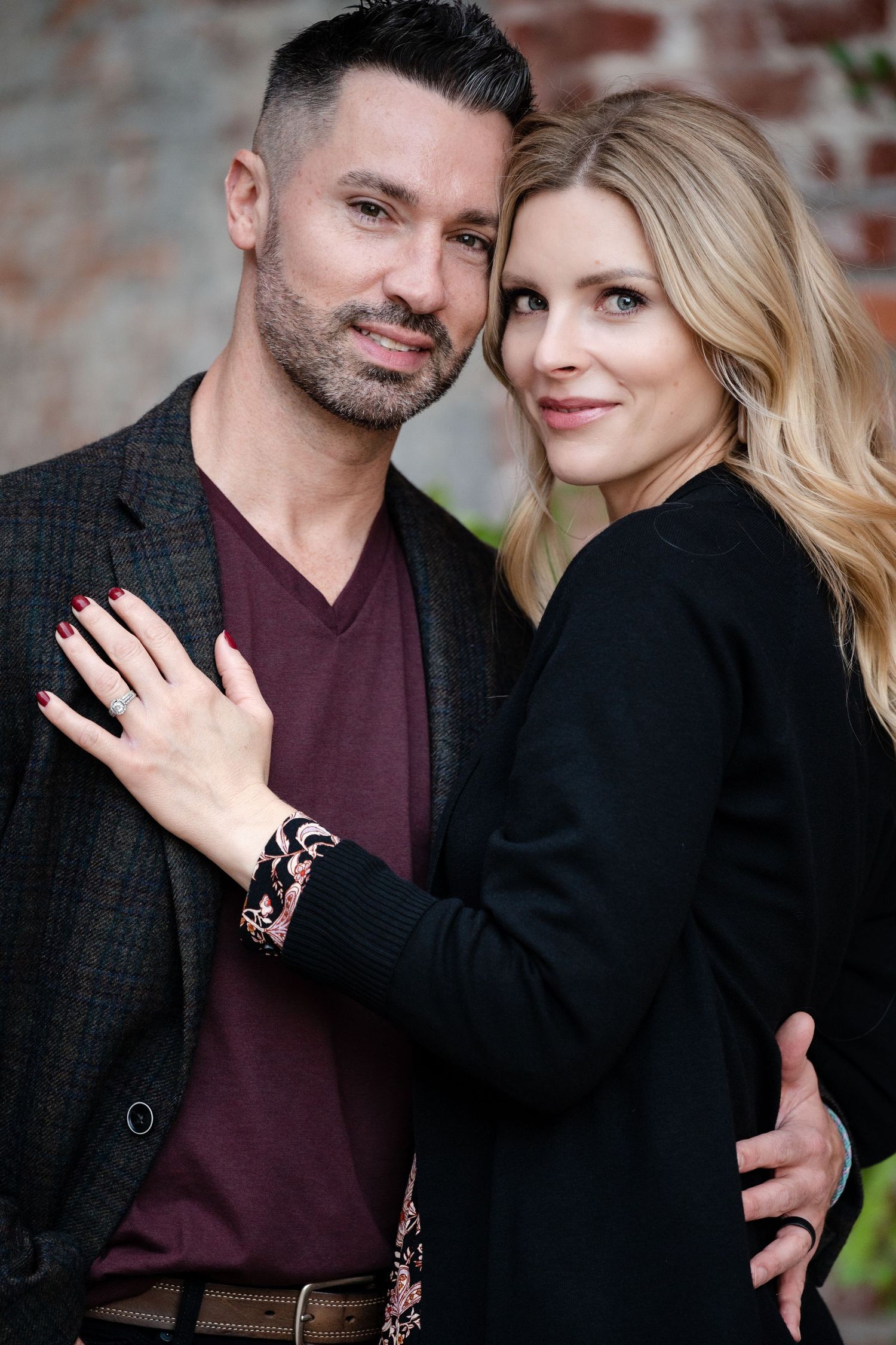 A stylish couple poses against a brick wall wearing complementary dark colored outfits.