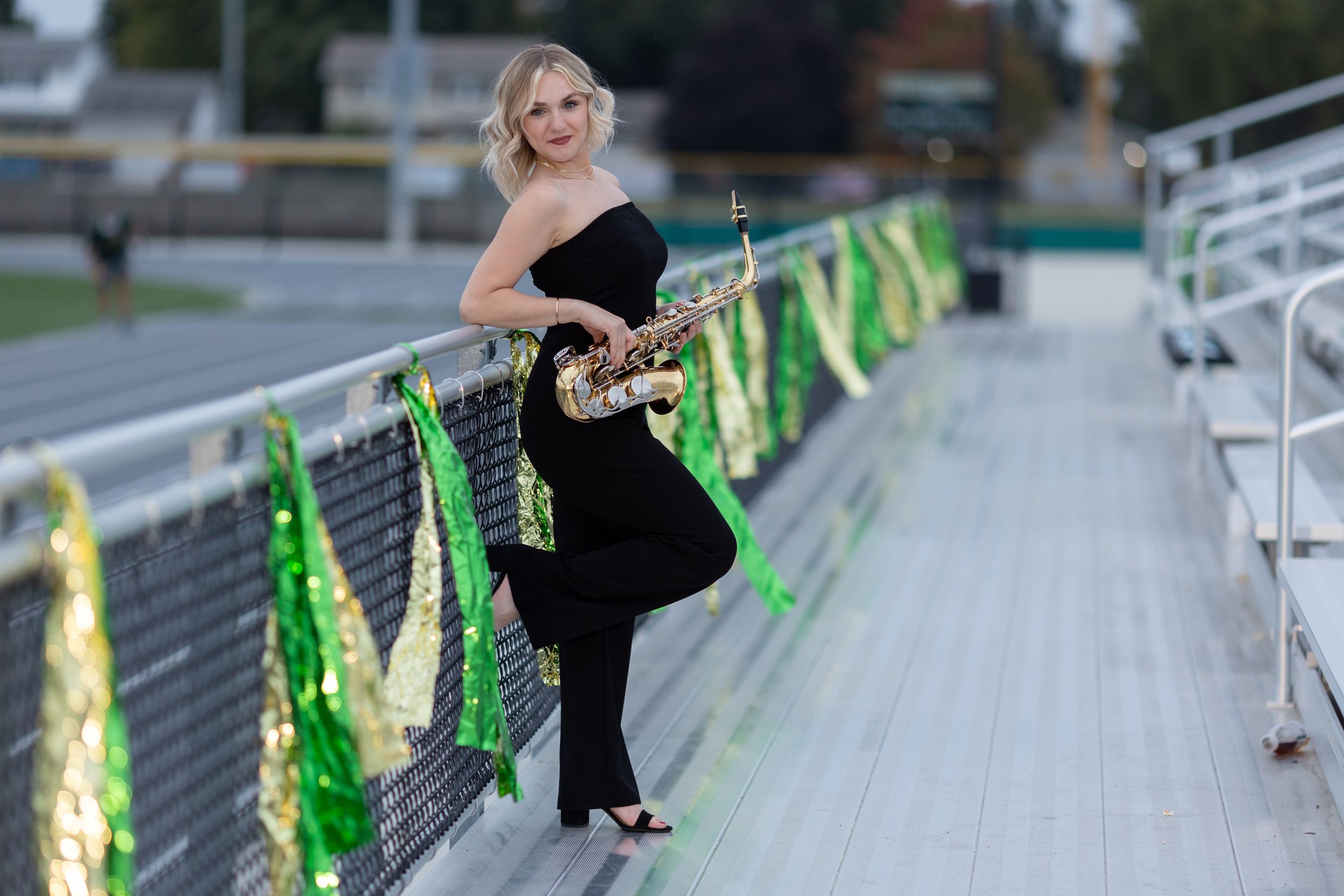 High school band participant leans against chain link fence with her saxophone.