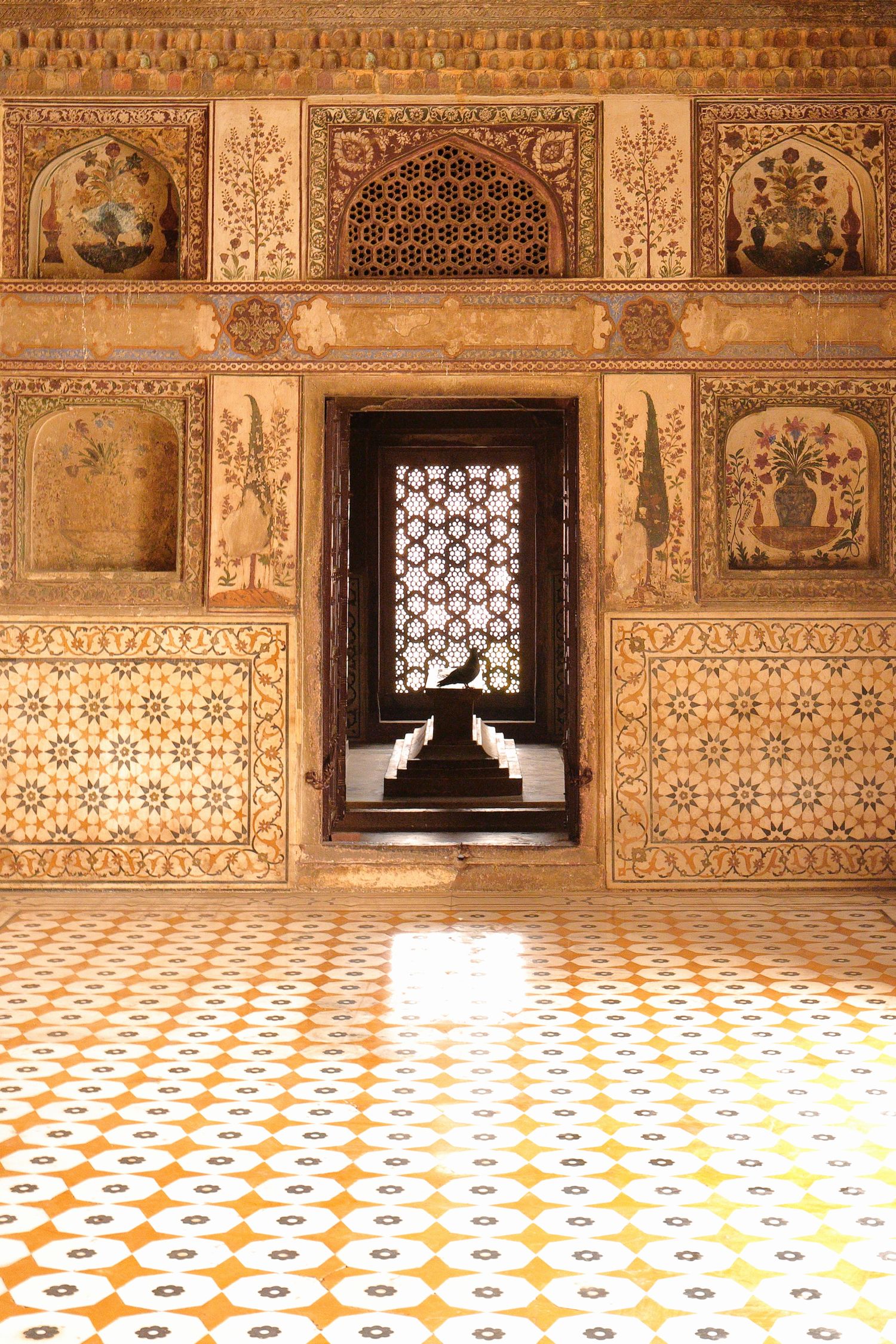 Ornate Mughal interior with intricate wall carvings, painted panels and geometric floor patterns in amber tones.