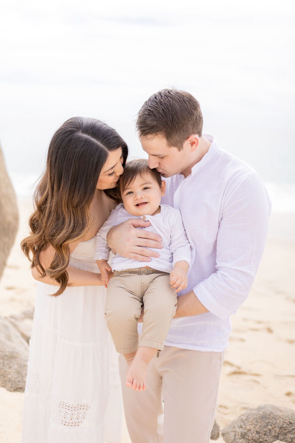 Family in neutral colored outfits sharing tender moments on sunny beach.