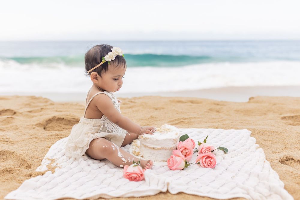 Baby sitting on floral blanket at beach with coral pink flowers and ocean waves.