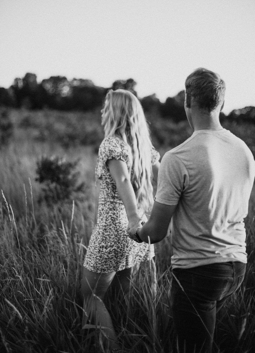 A romantic couple holding hands while standing in a field of tall grass at sunset, captured in black and white.
