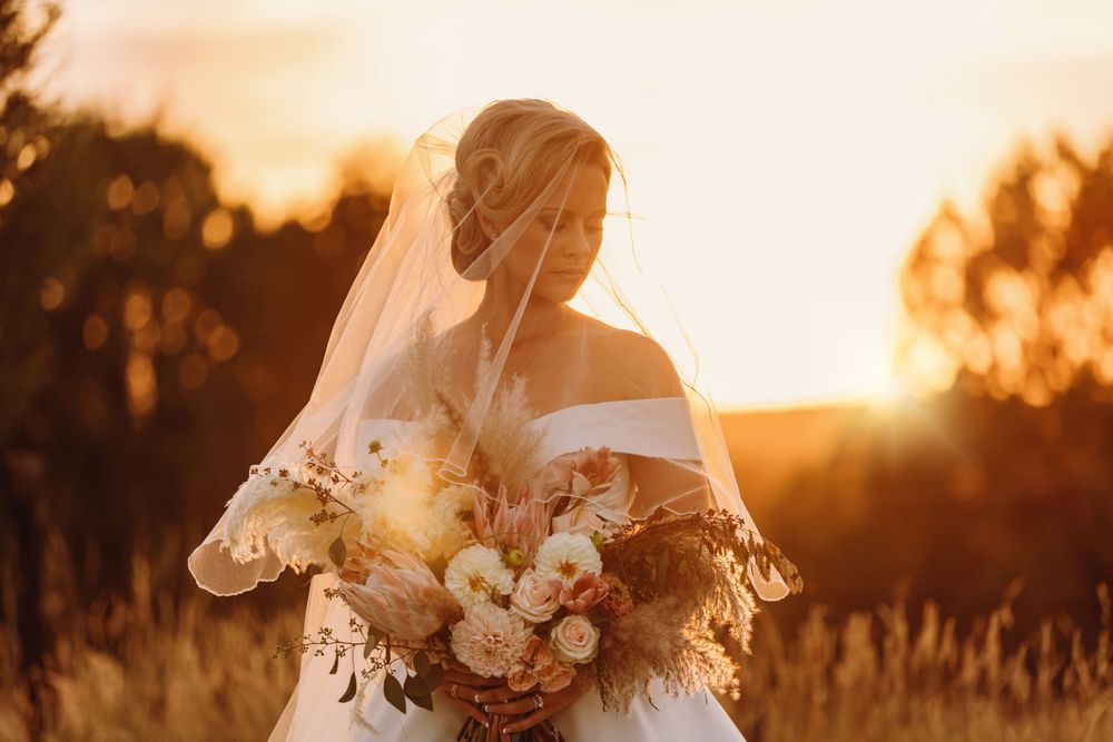Bride in white gown and veil holding bouquet of flowers in golden sunset field.