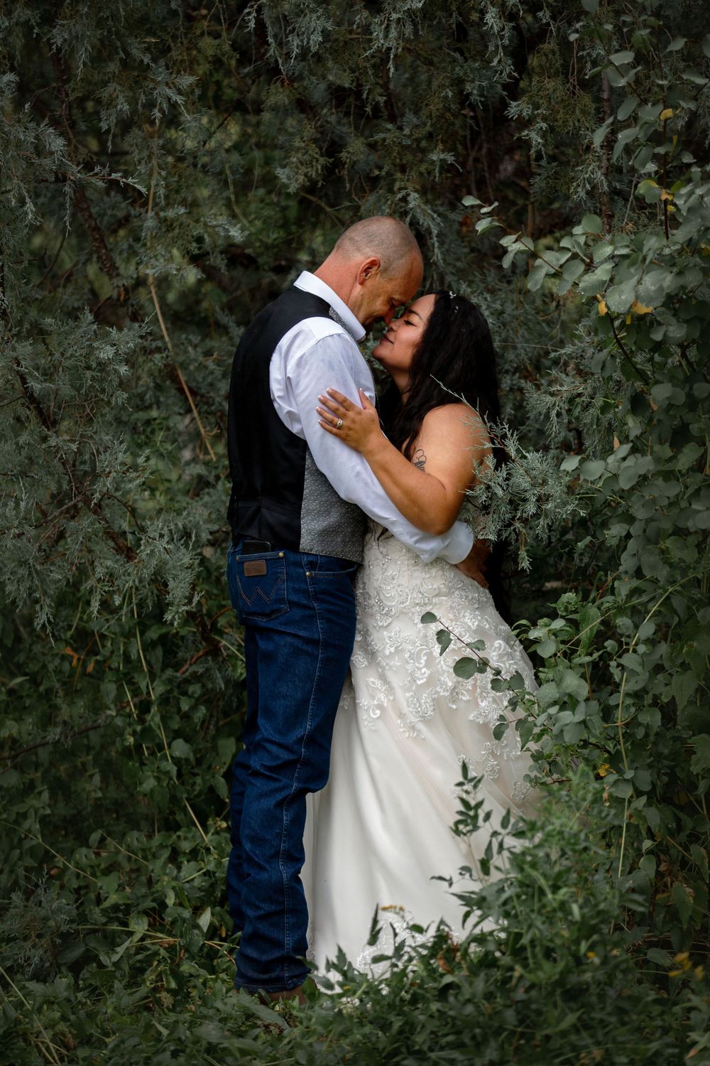 Couple shares a romantic kiss surrounded by lush green foliage, bride in lace gown and groom in vest.