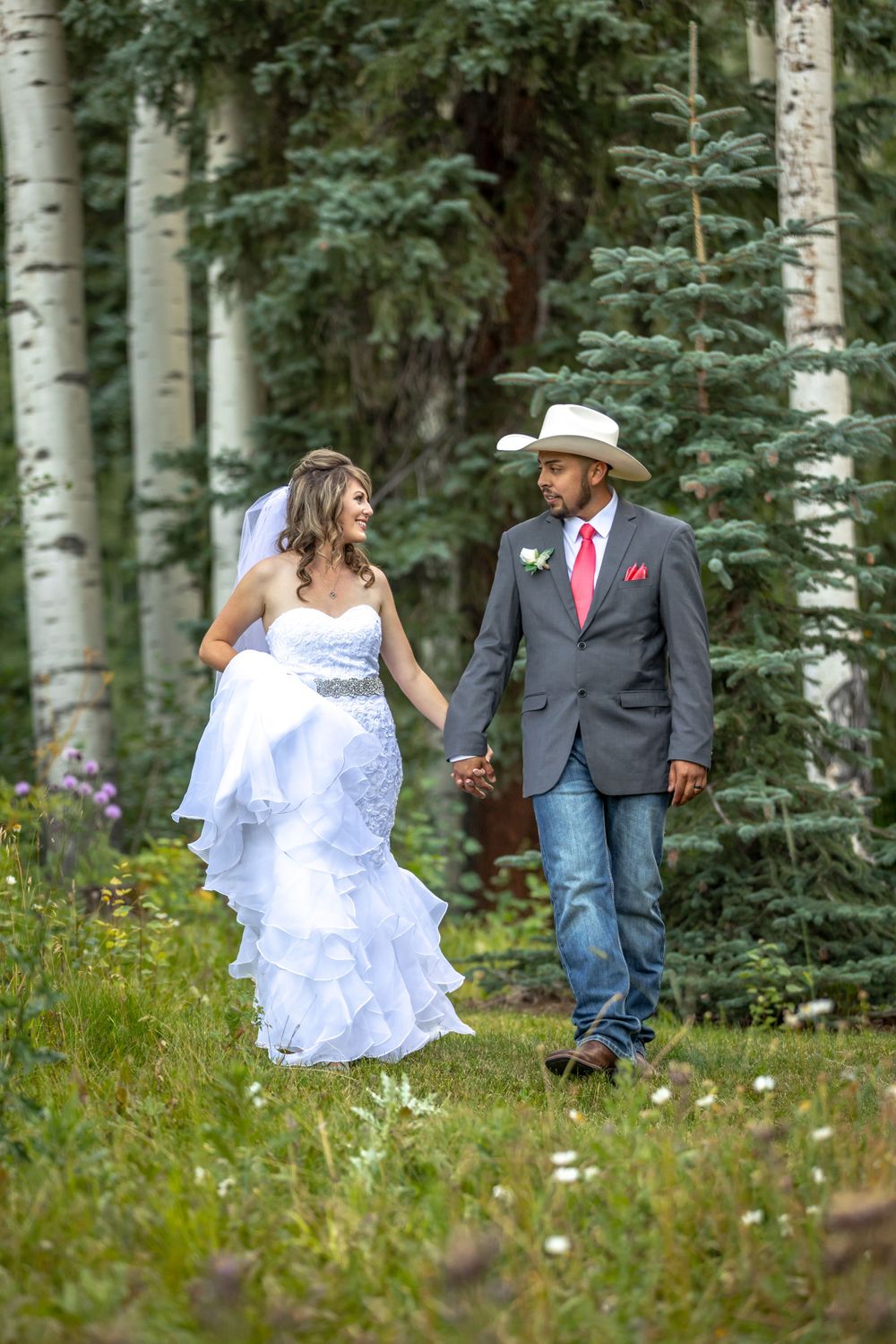 Bride and groom walking hand in hand through aspen forest in rustic western wedding attire.