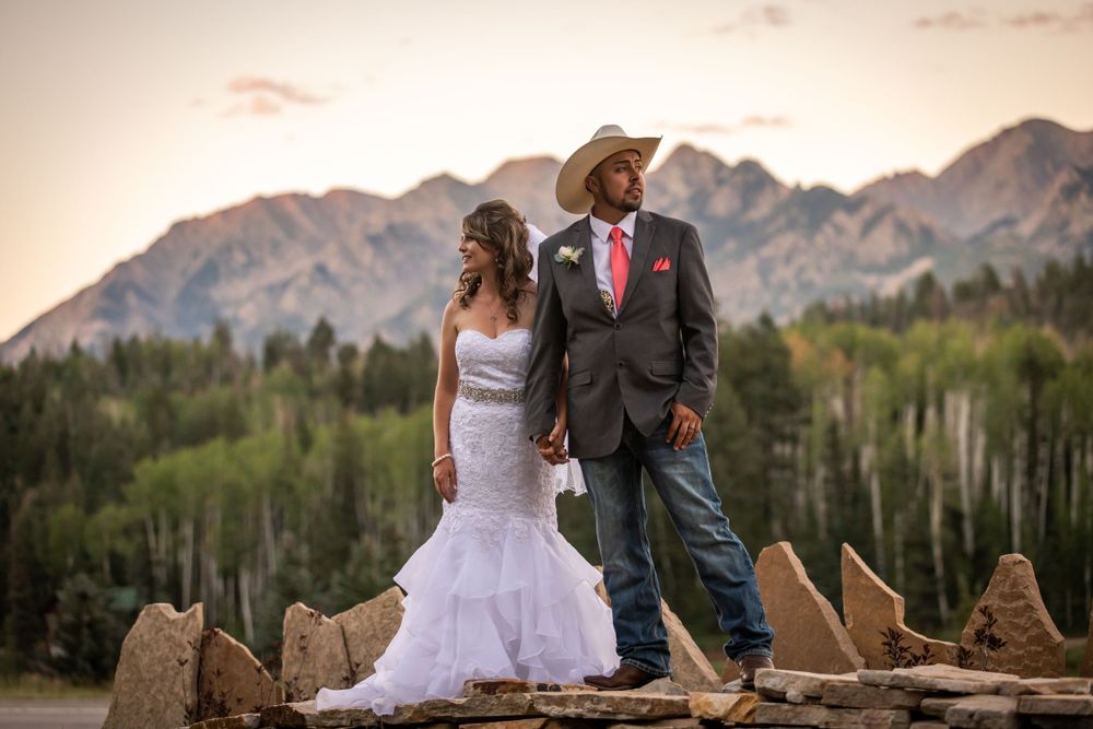 Bride and groom in western attire pose on rocks with mountain backdrop at sunset.