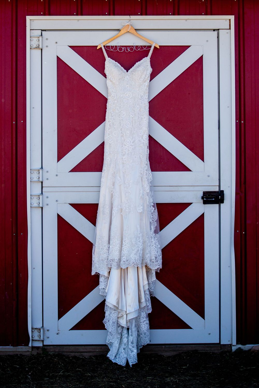 Elegant lace wedding dress hangs on a wooden hanger against a rustic red barn door.