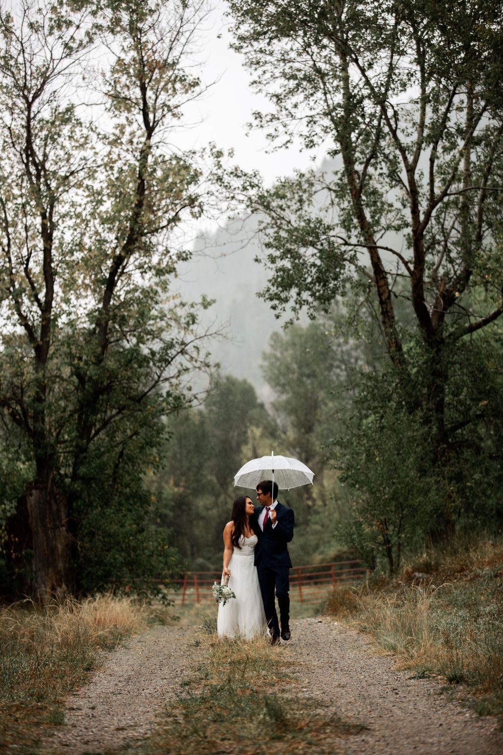 Bride and groom walk a forest path sharing a white umbrella on a misty wedding day.