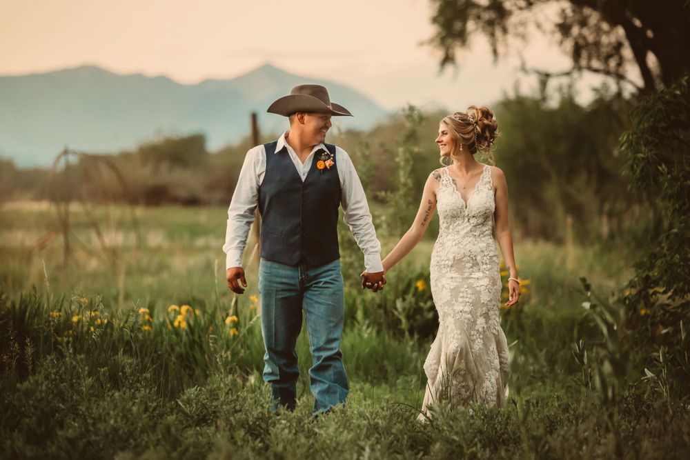 Couple in western wedding attire walk hand-in-hand through a wildflower meadow at sunset.