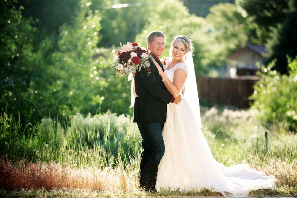Bride and groom pose together in a sunlit meadow, she holding a burgundy bouquet in her white gown.