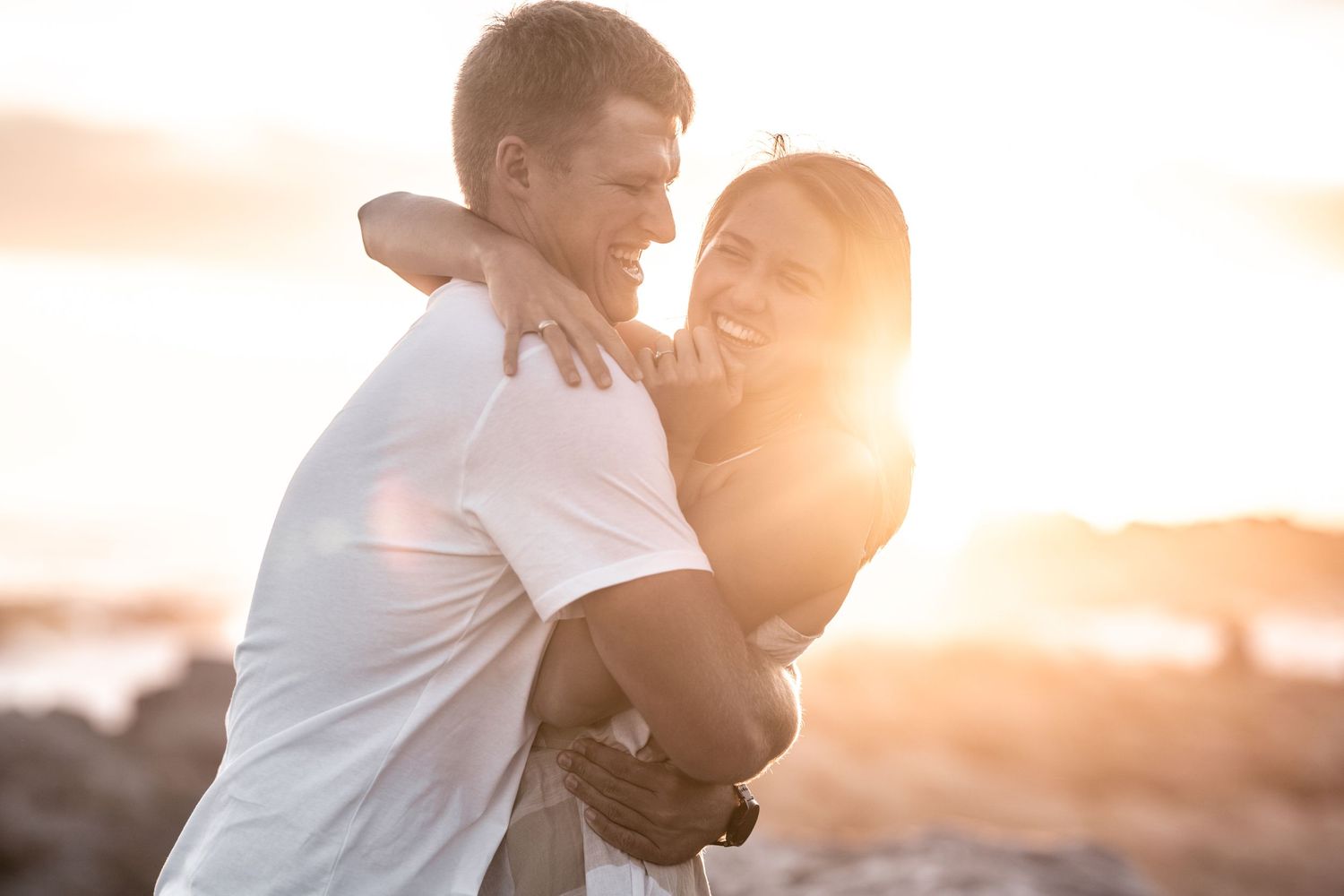 Joyful couple embraces and laughs together on a beach during a golden sunset engagement photoshoot.