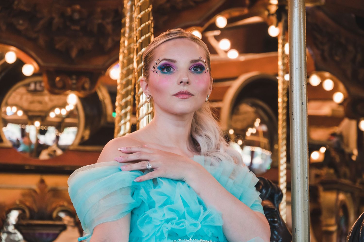 Blonde woman in teal dress with glitter eye makeup poses at ornate carousel.