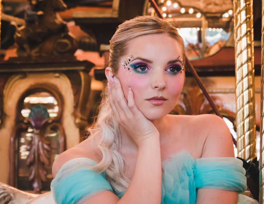 Young blonde woman with jeweled eye makeup in blue gown poses at carousel.