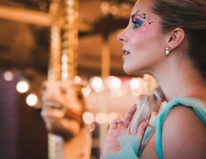 Side profile of woman with colorful glitter eye makeup in teal dress at carousel.