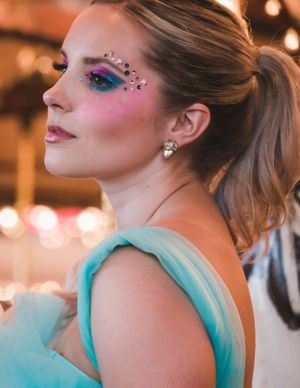 Woman with vibrant rainbow eye makeup and gems in teal dress at carousel.
