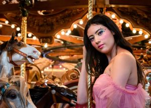 Dark-haired woman in pink dress with artistic eye makeup poses at carousel.