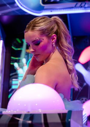 Blonde woman with purple gem eye makeup in white dress at neon arcade.