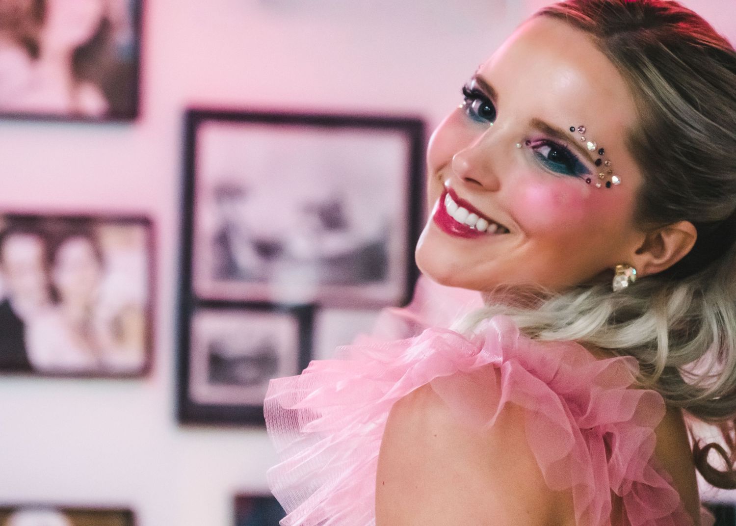 Smiling woman with pink eye makeup and gems in pink tulle dress indoors.