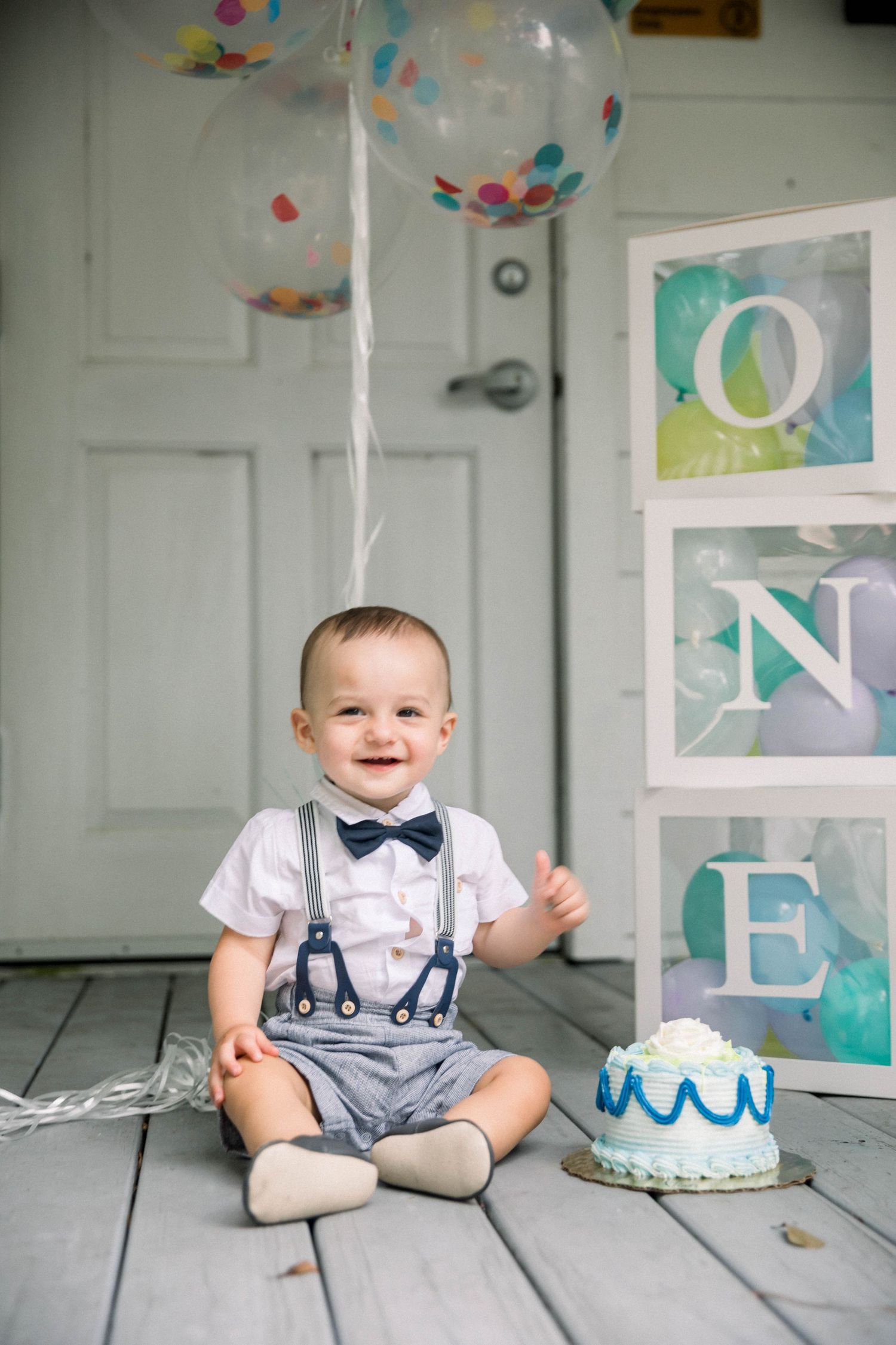A cake smash photo session featuring a one year old in suspenders and bow tie sitting next to a white birthday cake.
