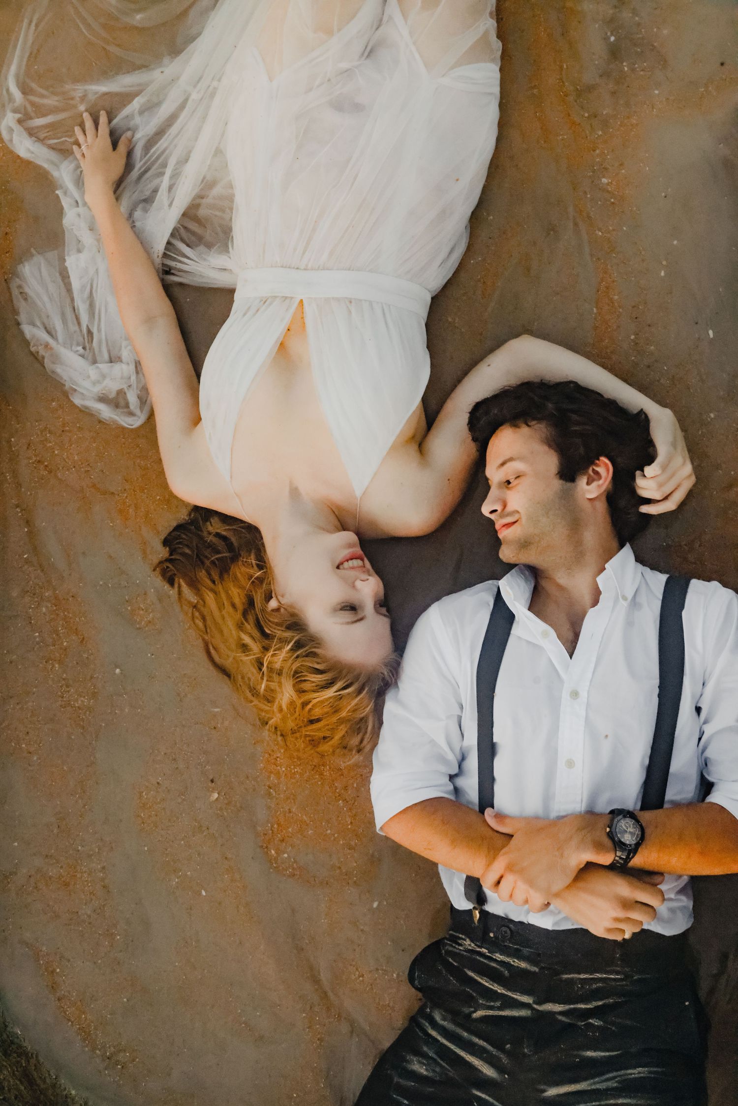 Artistic couple's photo showing contrasting poses on a textured wall with suspenders and flowing white dress.