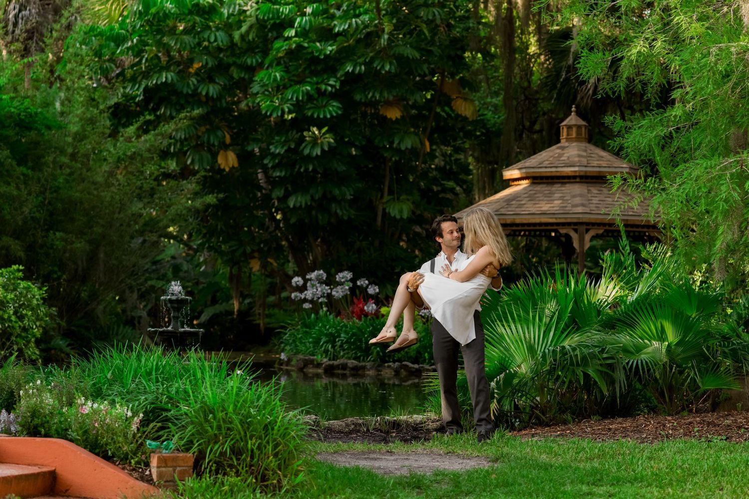 Couple poses dramatically in a lush garden setting with a gazebo.