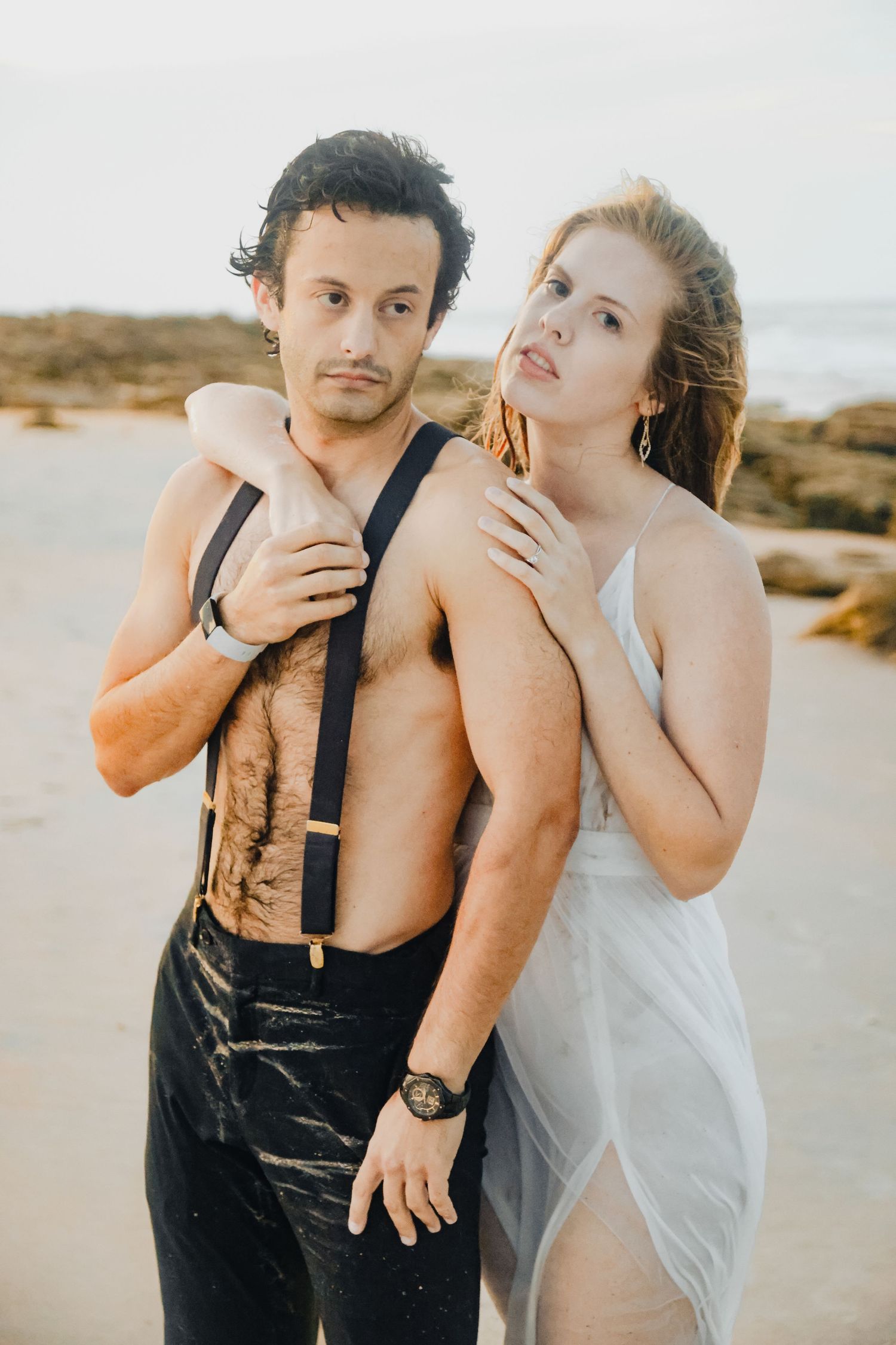 Romantic beach photoshoot featuring couple in white dress and black suspenders against coastal backdrop.