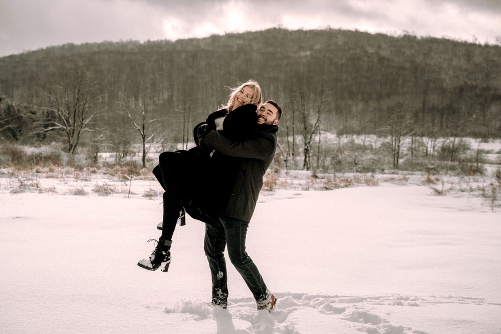 A playful couple spinning in the snow with mountains and cloudy skies in the background.
