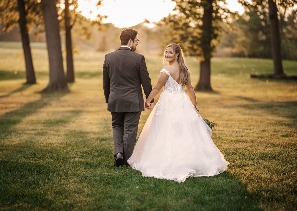Romantic sunset wedding portrait of couple walking through tree-lined field.