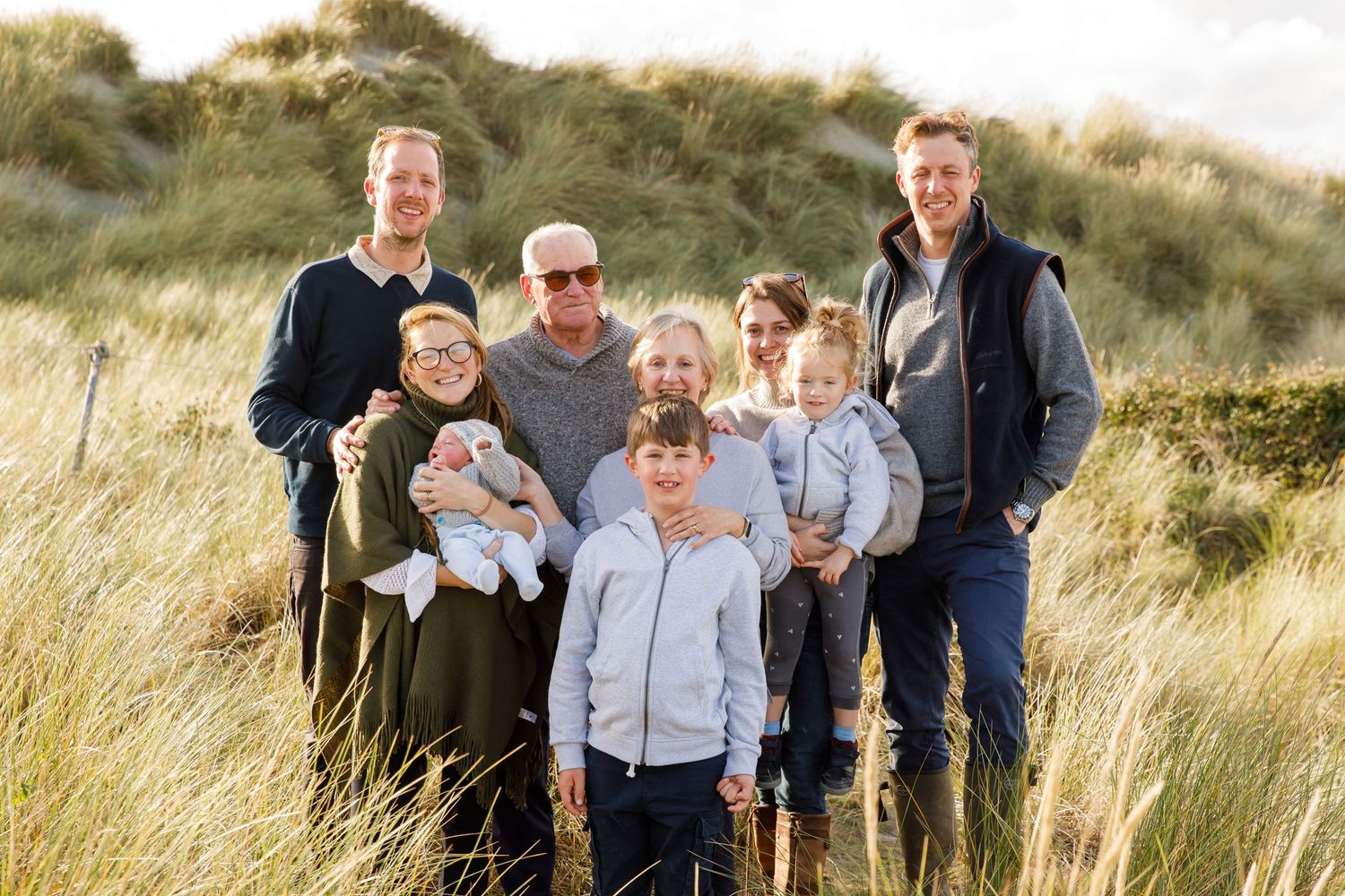 A multi-generational family group poses together among coastal dune grass during a family beach photoshoot