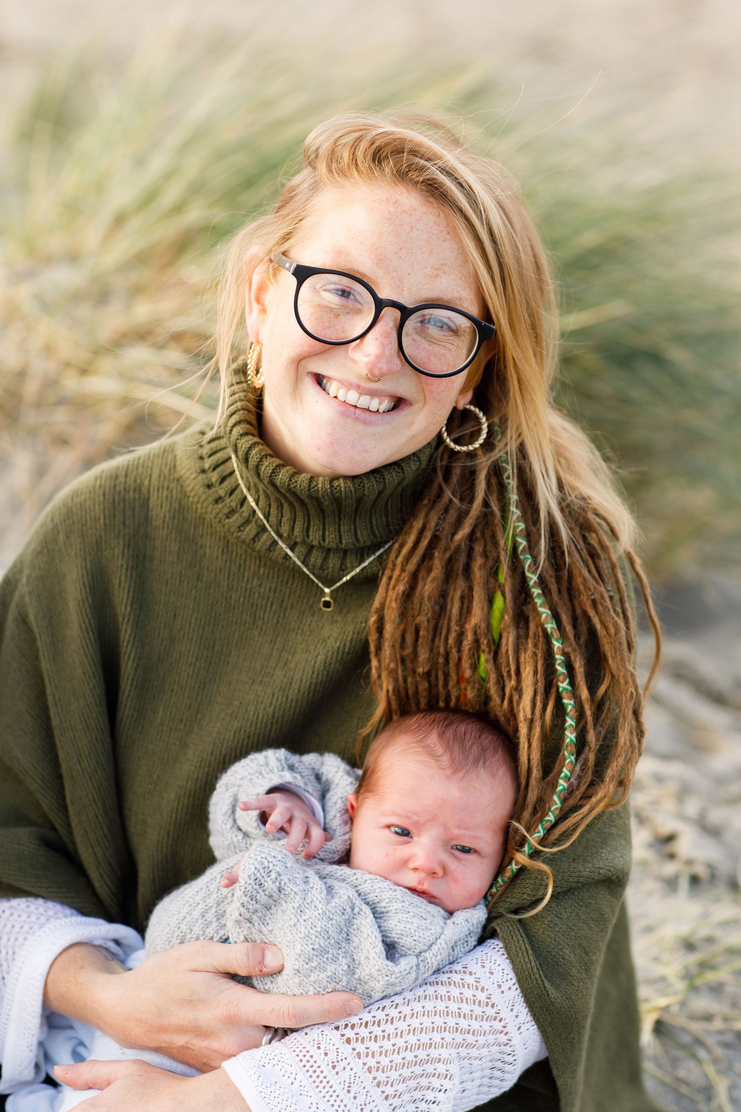 Baby in a cozy gray sweater being held and smiling with an adult wearing glasses and green knit sweater during a family beach photoshoot