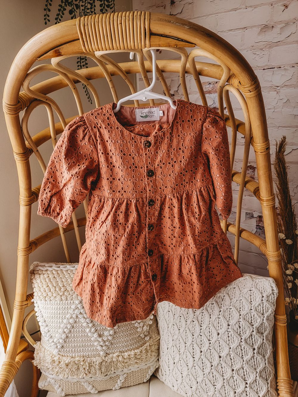 Rust brown eyelet lace short sleeve toddler dress with button front hanging on rattan chair.