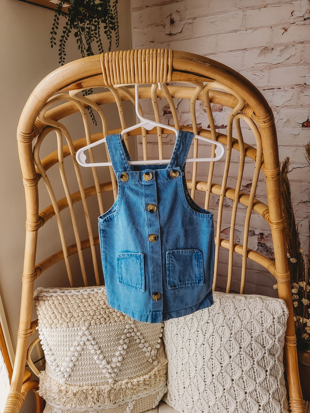 Small denim button-front pinafore dress with pockets hanging on white hanger on rattan chair.